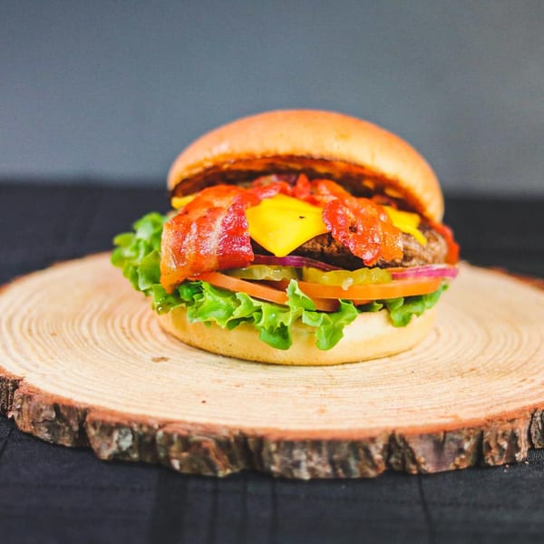 Bacon cheeseburger with lettuce, tomato, onion, and pickles on a wooden slab.